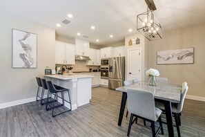 A modern kitchen with white cabinetry, a central island with bar stools, stainless steel appliances, and a dining area with a marble table. - 10 Mi to Disney & Old Town! Stylish Family Home (Kissimmee)