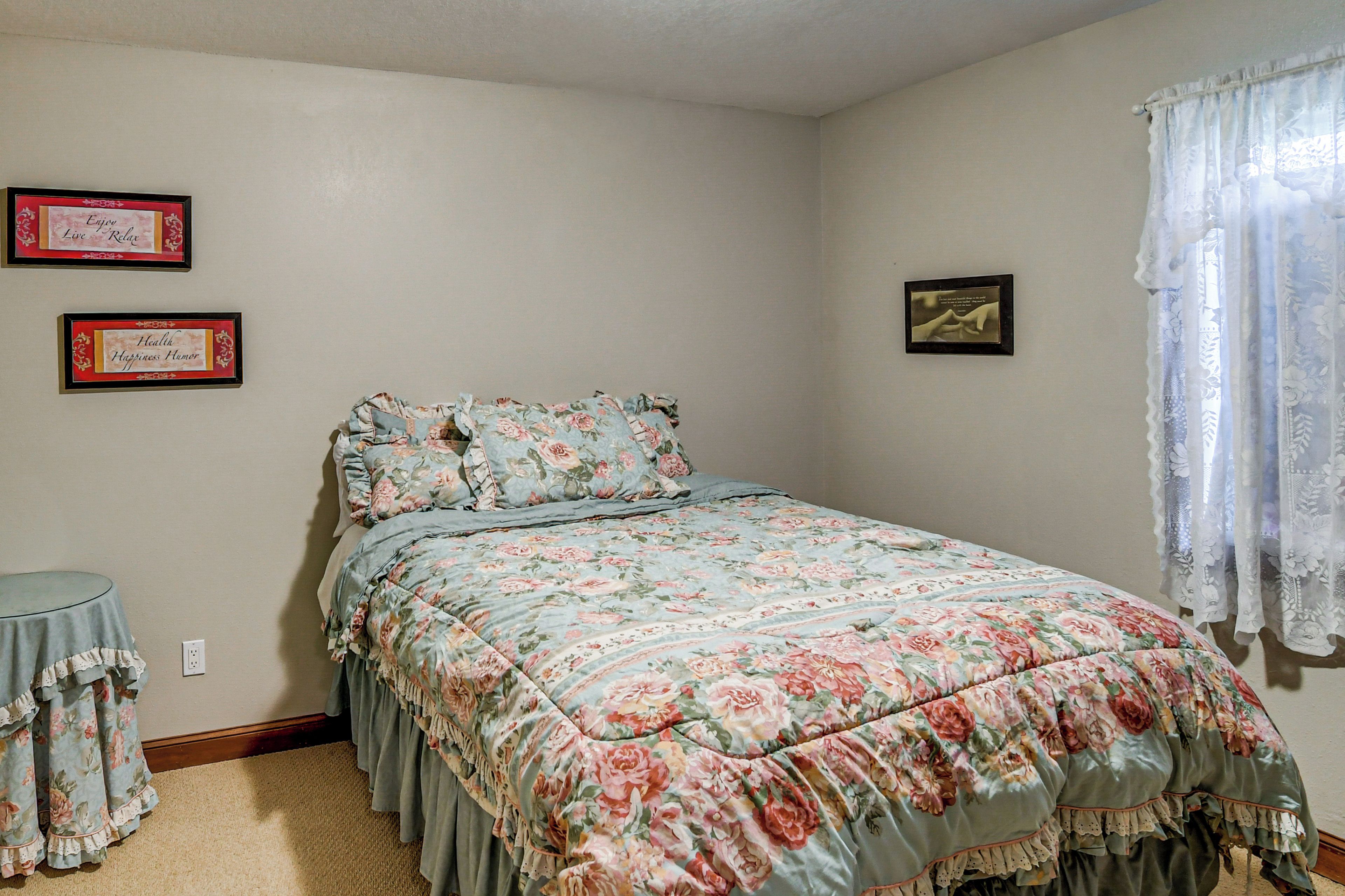 A bedroom with a floral bedspread, a small round table, and two framed items on the wall.