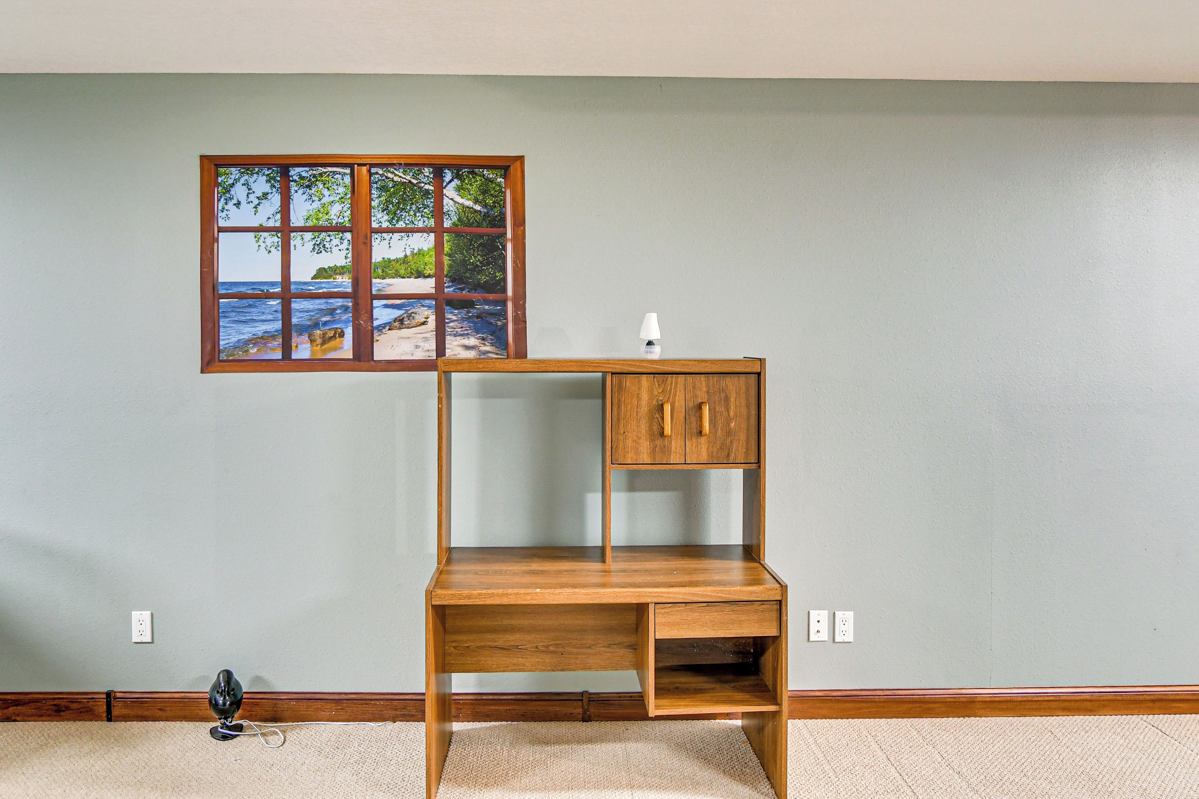 A room with a wooden desk against a wall, a window with a view of the beach, and a lamp on the desk.