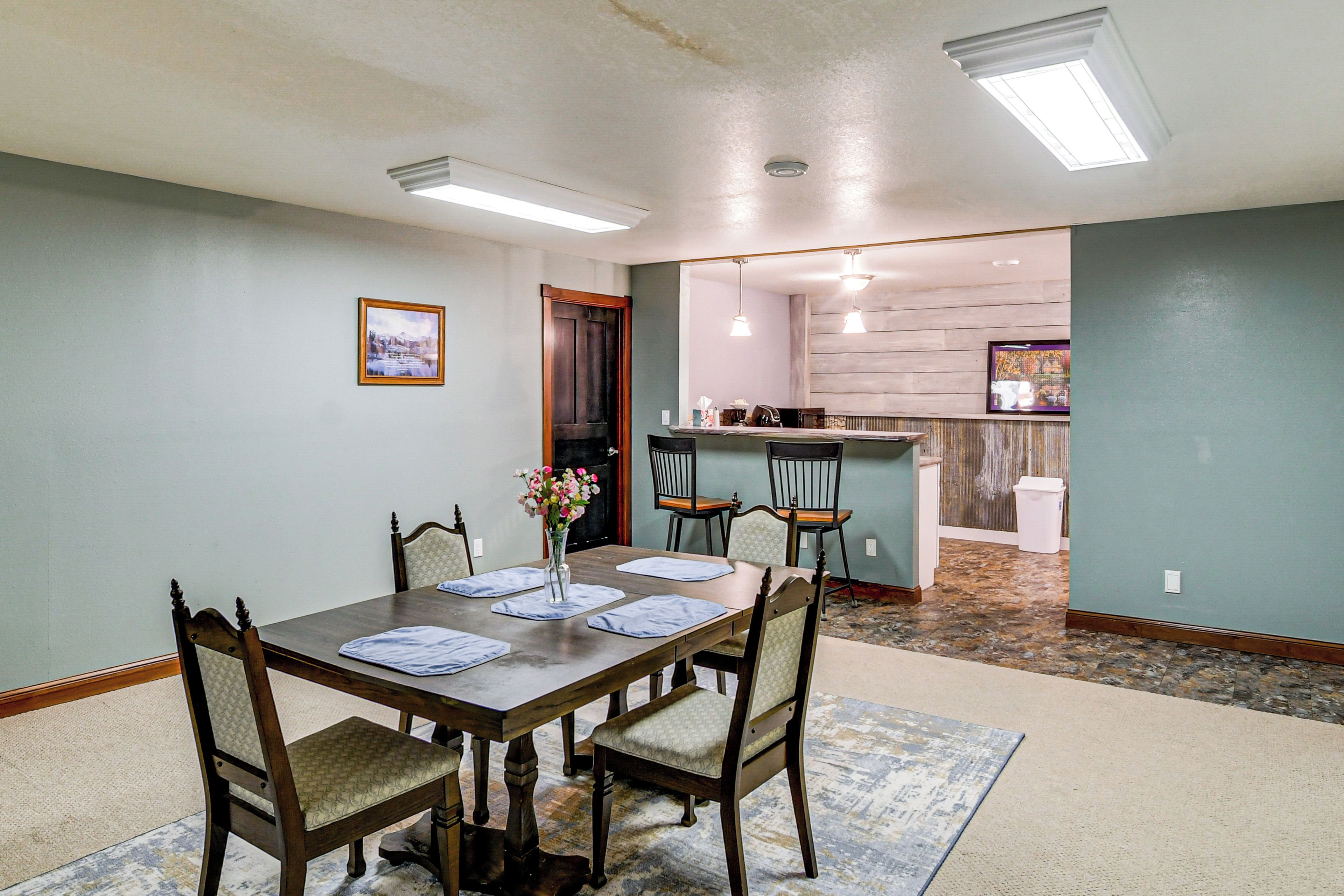 A dining area with a wooden table, four chairs, and a vase of flowers. There is a kitchen in the background with a bar and a trash can.