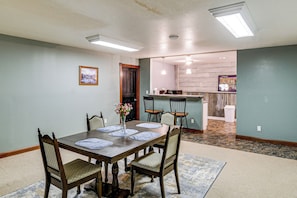 A dining area with a wooden table, four chairs, and a vase of flowers. There is a kitchen in the background with a bar and a trash can.