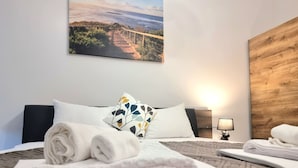 Une chambre d’hôtel avec un lit, une table de chevet avec une lampe, une tête de lit en bois et un tableau représentant un paysage côtier.