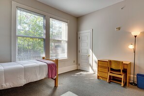 Un dormitorio con cama, escritorio y silla. Hay una ventana con persianas, una puerta y una lámpara de pie.