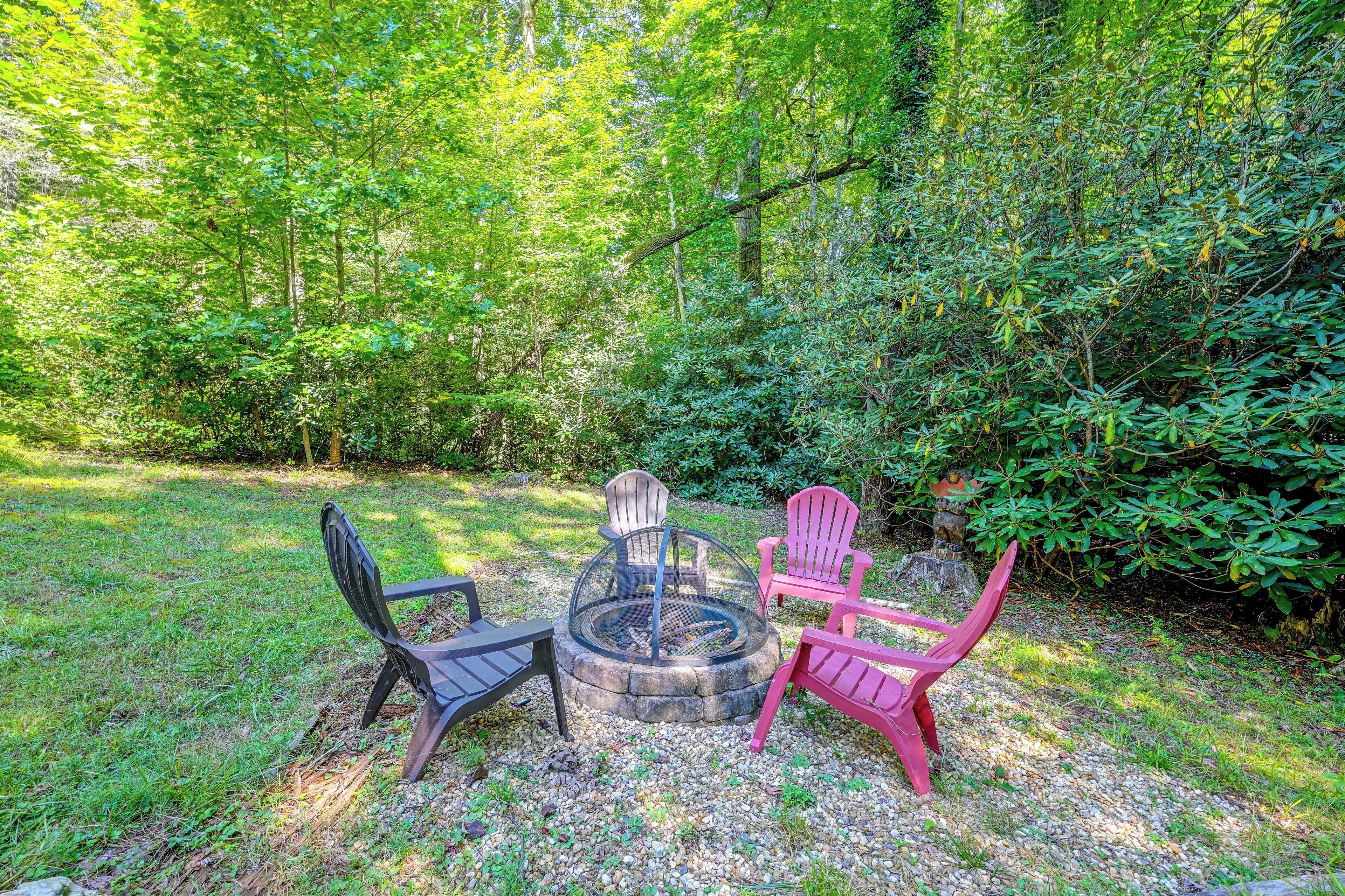 A circular fire pit with four chairs around it, set on a gravel area with grass and trees in the background.