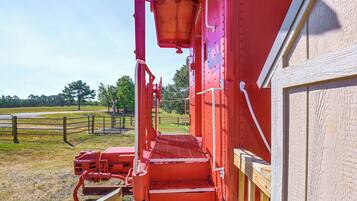 Un wagon de train rouge, avec un escalier métallique menant à un quai, entouré d’une clôture en bois et d’arbres.