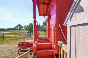 Ein roter Zugwaggon mit einer Metalltreppe, die zu einer Plattform führt, umgeben von einem Holzzaun und Bäumen.