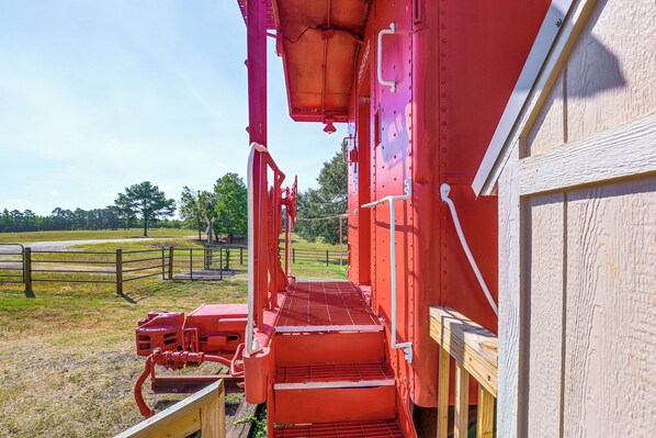 A red train car with a metal staircase leading to a platform, surrounded by a wooden fence and trees.