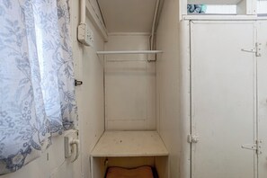 Ein schmales Zimmer mit einem Einzelbett, einer kleinen Ablagefläche und einer Tür.