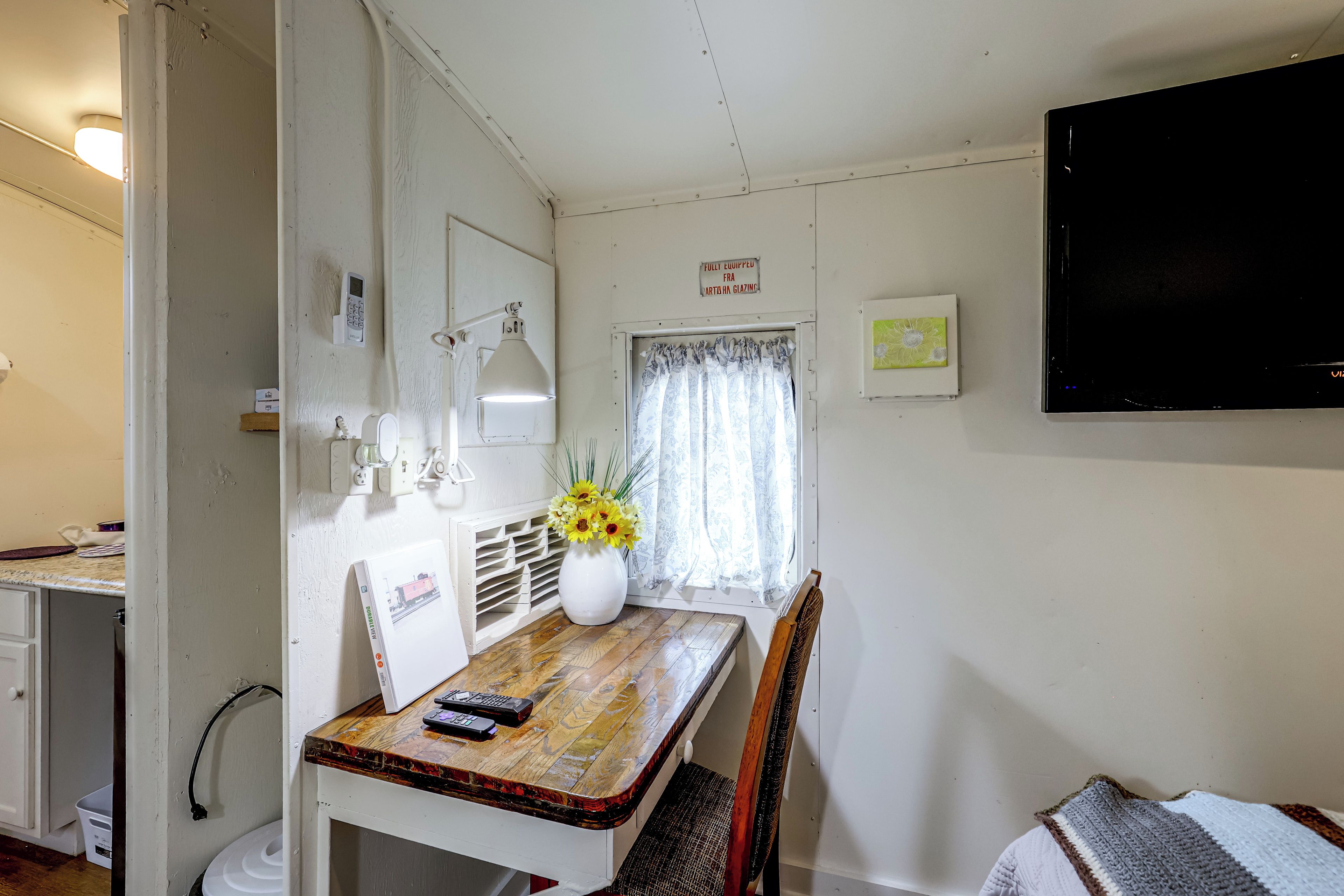 Une petite pièce, de taille modeste, avec une table en bois, une chaise, un bureau avec un ordinateur, un téléviseur fixé au mur et une fenêtre avec des rideaux.