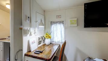 Une petite pièce, de taille modeste, avec une table en bois, une chaise, un bureau avec un ordinateur, un téléviseur fixé au mur et une fenêtre avec des rideaux.