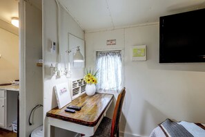 A small, compact room with a wooden table, a chair, a desk with a computer, a wall-mounted TV, and a window with curtains.