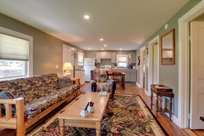 A living room with a sofa, a coffee table, and a patterned rug. There is a kitchen area with cabinets and a refrigerator.
