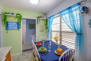 Un comedor con una mesa dispuesta para cuatro personas, una ventana con cortinas azules y un cartel en la pared.