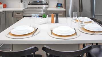 Una mesa de comedor dispuesta con platos blancos, cubiertos y vasos transparentes, con una pequeña planta en maceta y dos saleros y pimenteros sobre la mesa.