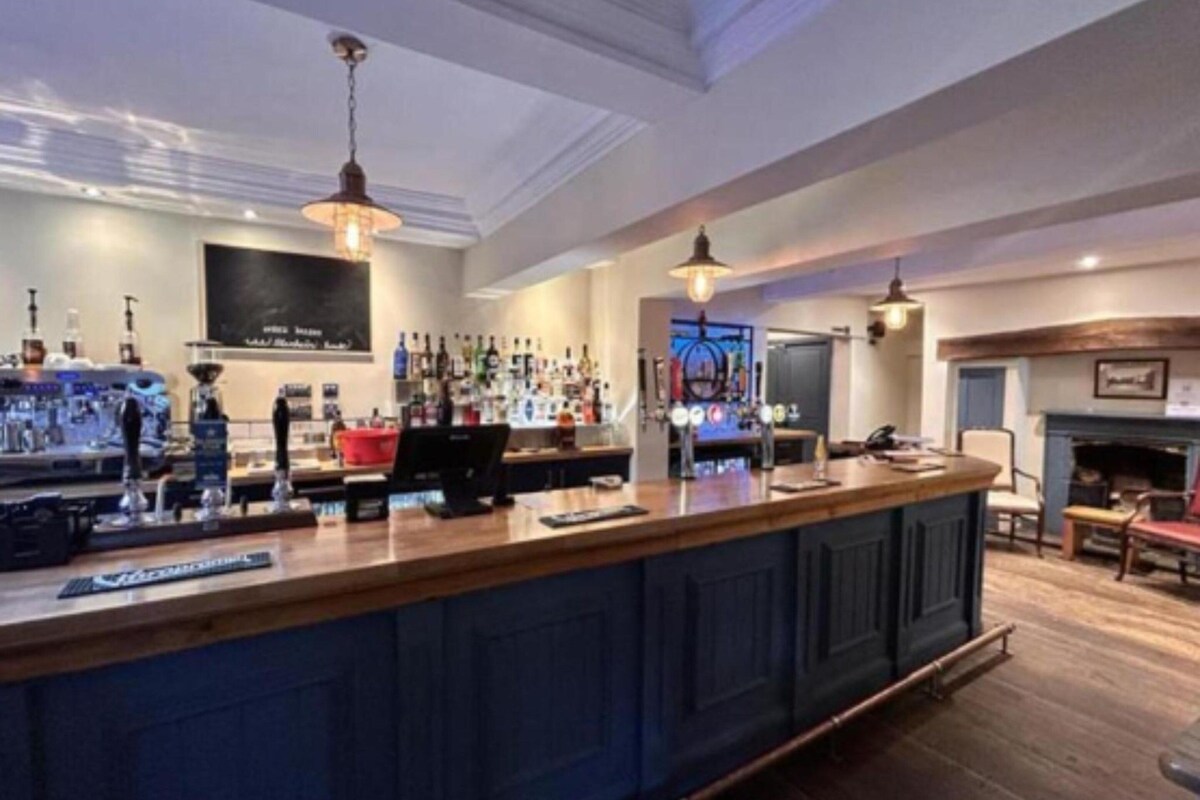 A bar area with a counter, shelves of bottles, and a fireplace in the background.