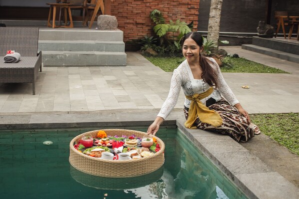 A person sitting by a pool with a basket of food and drinks.