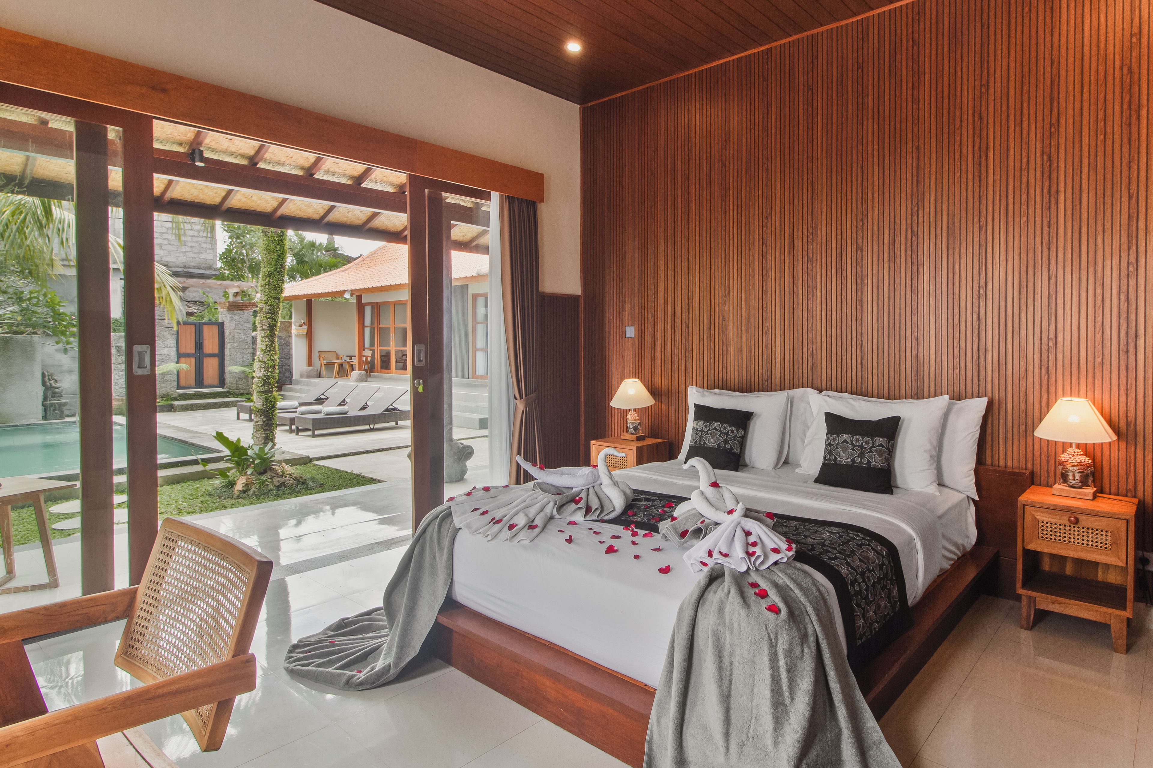 Habitación de hotel con cama, lámparas de noche, pared de madera y vista a un área de piscina exterior.