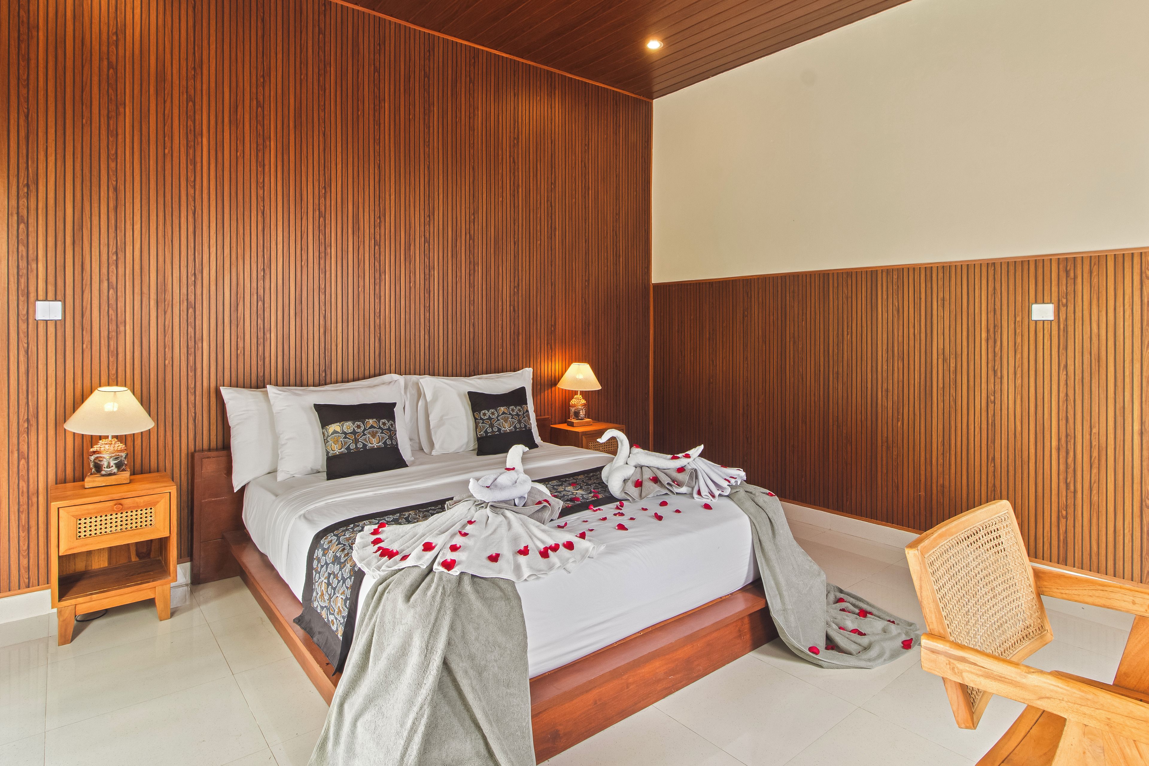 Habitación de hotel con pared de madera, una cama con ropa de cama blanca, dos lámparas de noche, una mesita de noche de madera y una silla de mimbre.