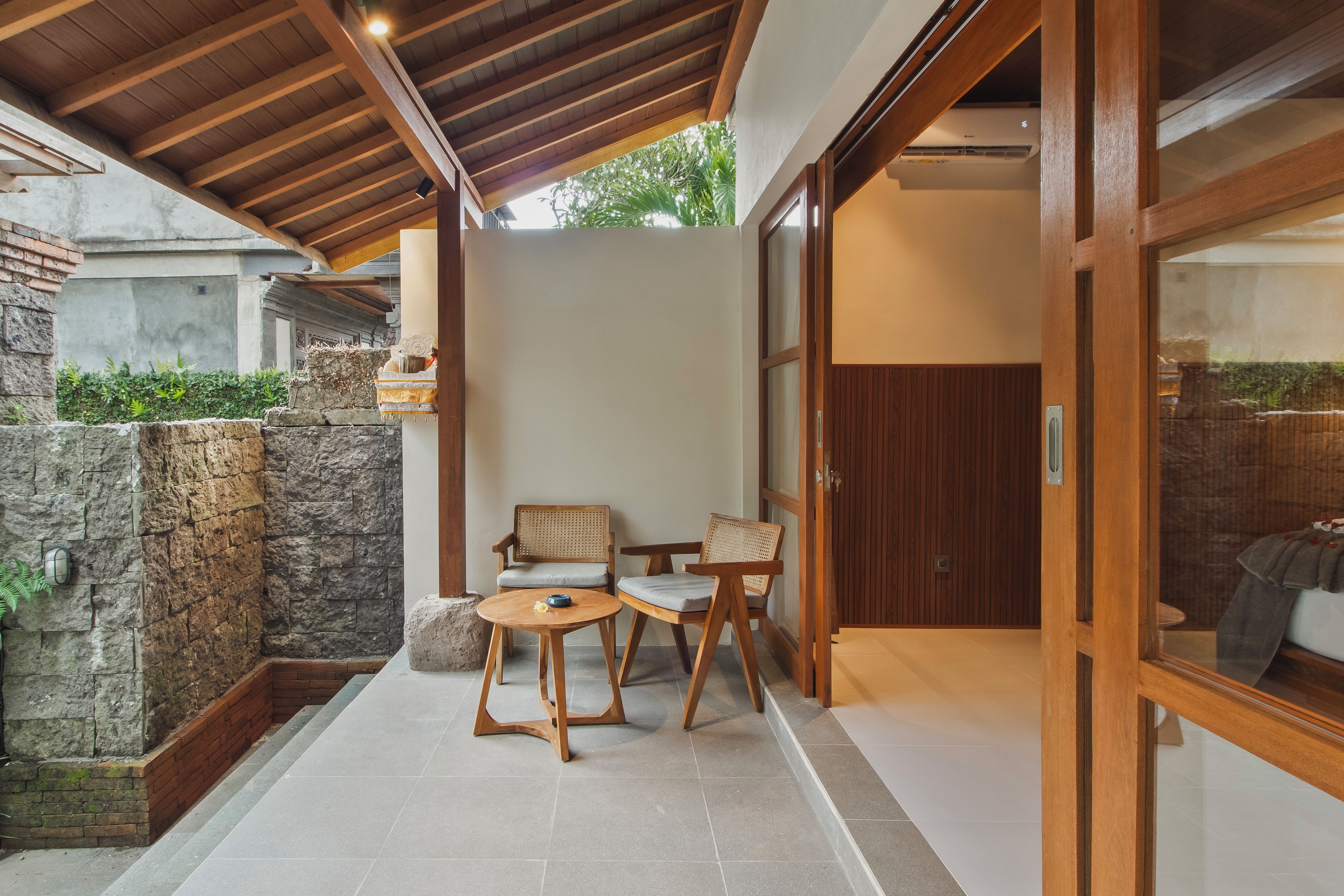 Un espacio moderno al aire libre con muros de piedra, mobiliario de madera y un ventanal que da a una habitación.