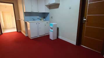 Une cuisine de taille réduite avec des meubles blancs, un évier et un réfrigérateur. On y trouve une porte avec un panneau indiquant un numéro, ainsi qu’un sol recouvert d’une moquette rouge.