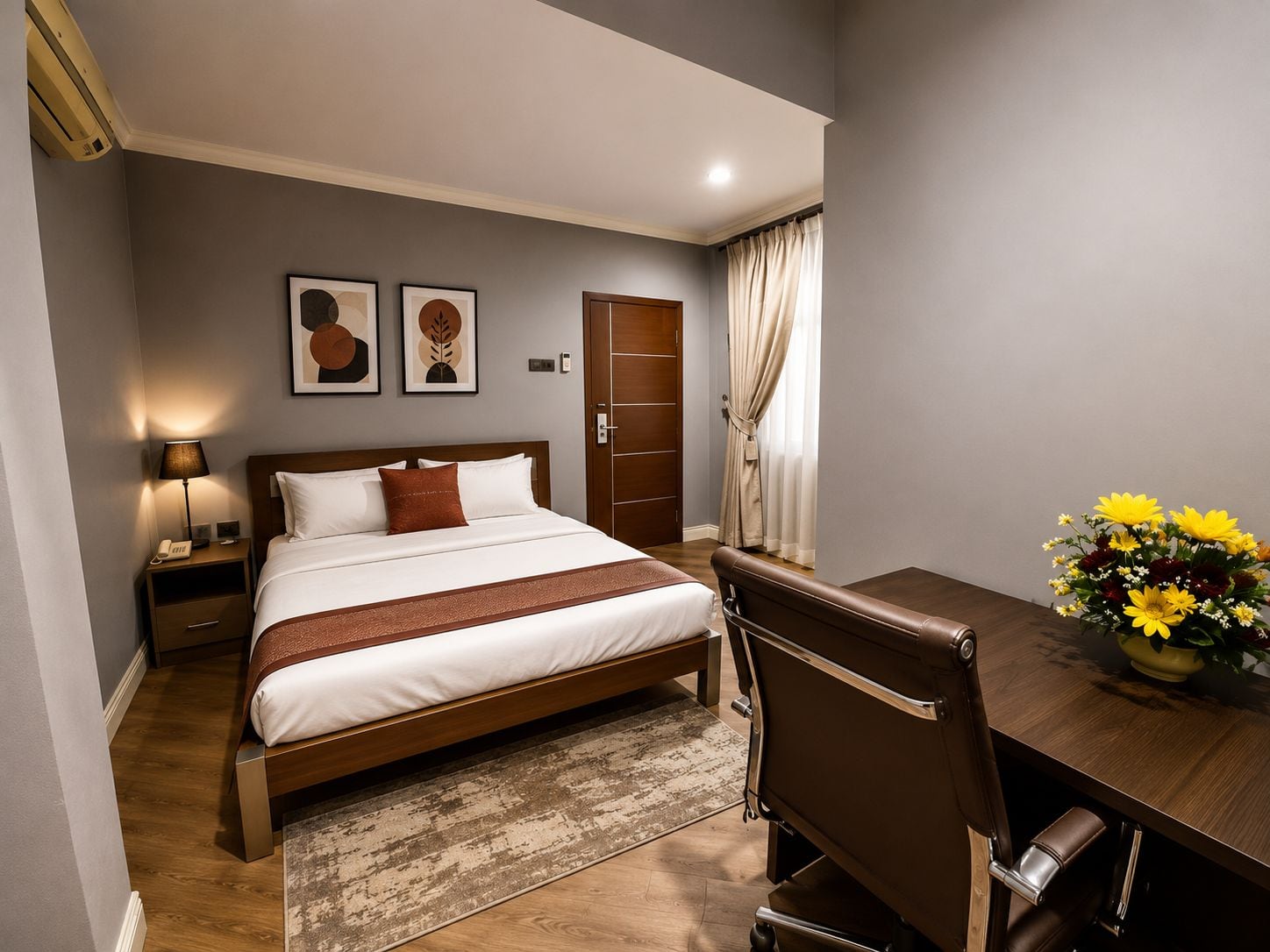 A hotel room with a bed, bedside table, lamp, artwork, a desk with a chair, and a vase of flowers.