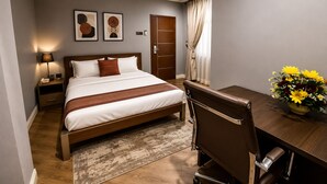 A hotel room with a bed, bedside table, lamp, artwork, a desk with a chair, and a vase of flowers. - Quiet Pols Residence  (Lagos)