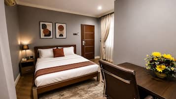 A hotel room with a bed, bedside table, lamp, artwork, a desk with a chair, and a vase of flowers.