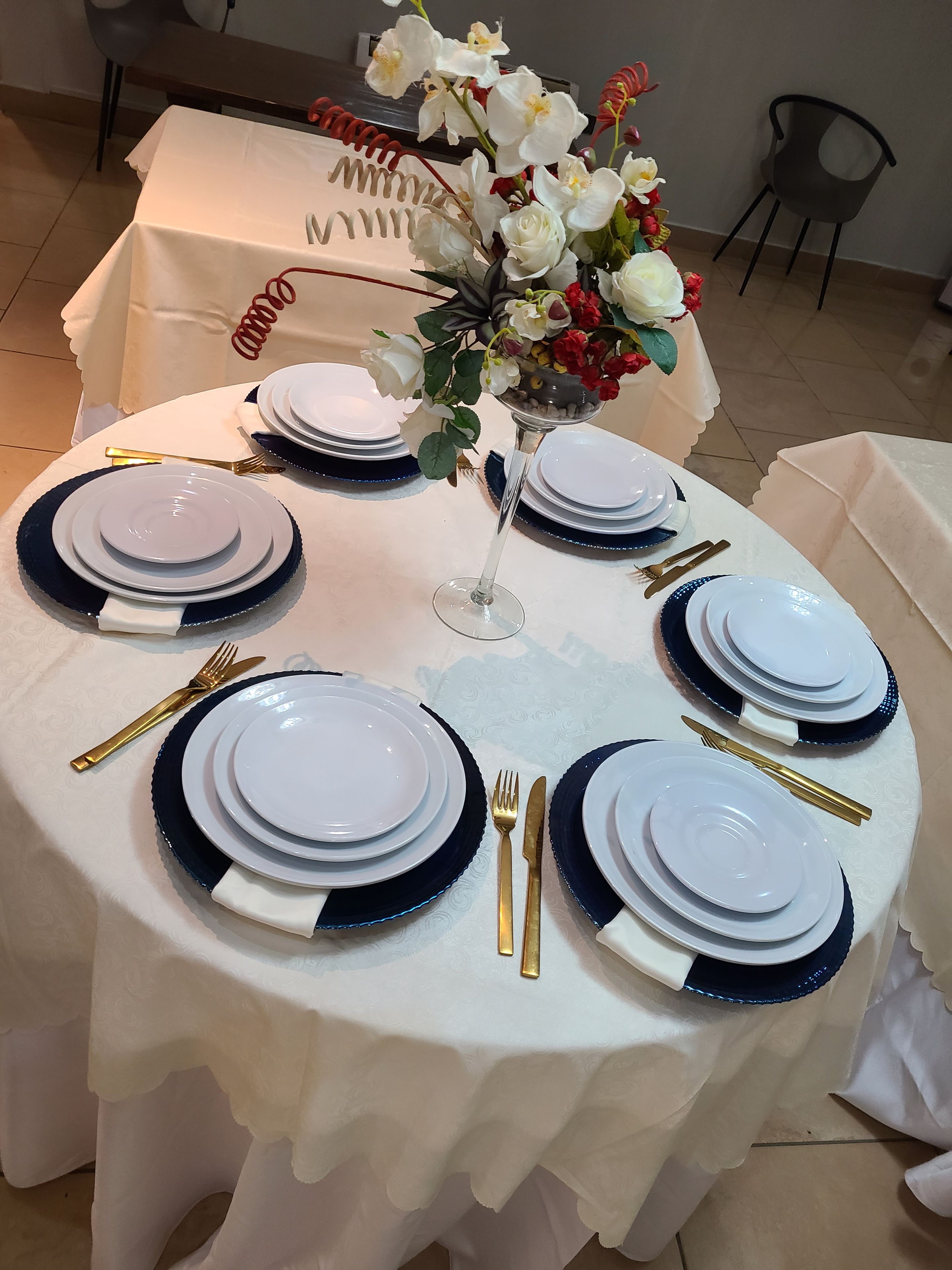 A round table set for a meal with white plates, blue rimmed plates, gold cutlery, and a vase with flowers.