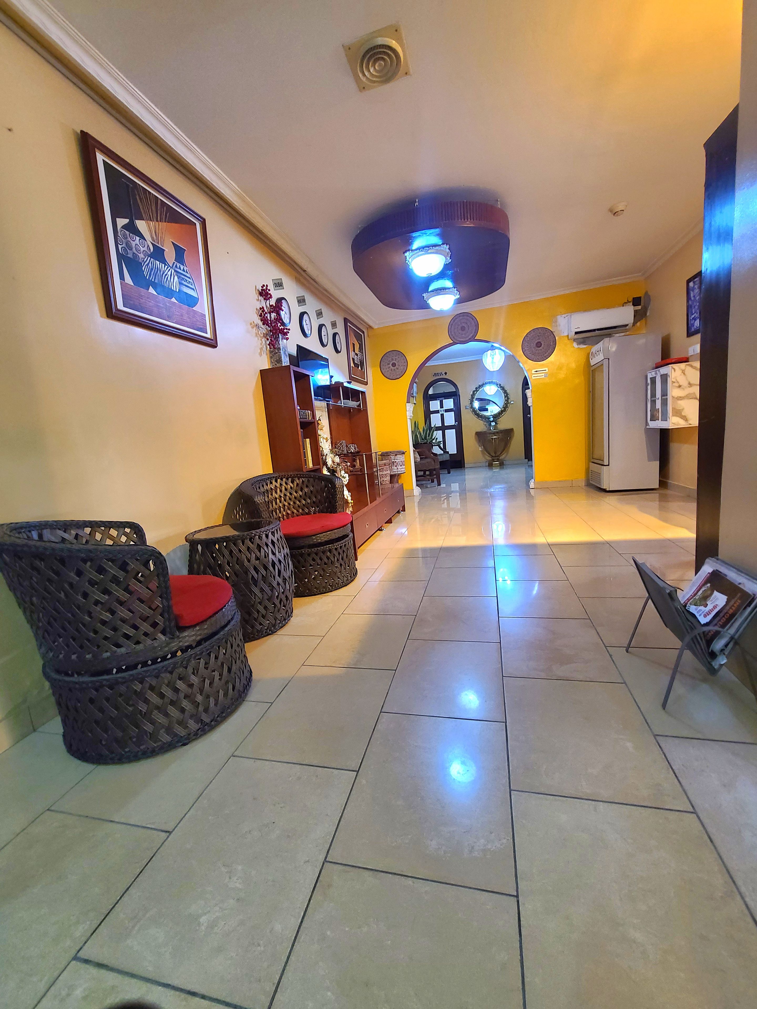 A hotel lobby with a reception desk, wicker chairs with red cushions, a wall-mounted clock, and a ceiling fan.