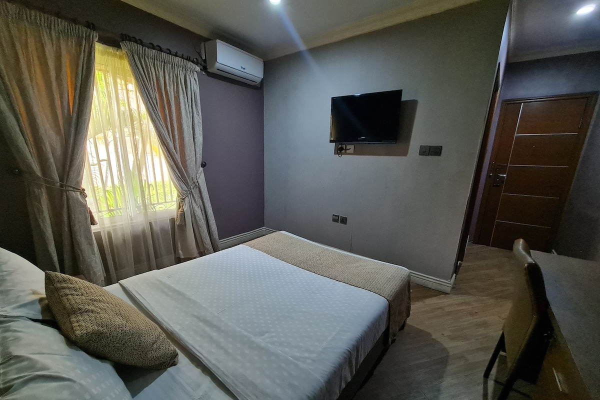 Quarto de hotel com cama, escrivaninha, ar condicionado, televisão e porta.