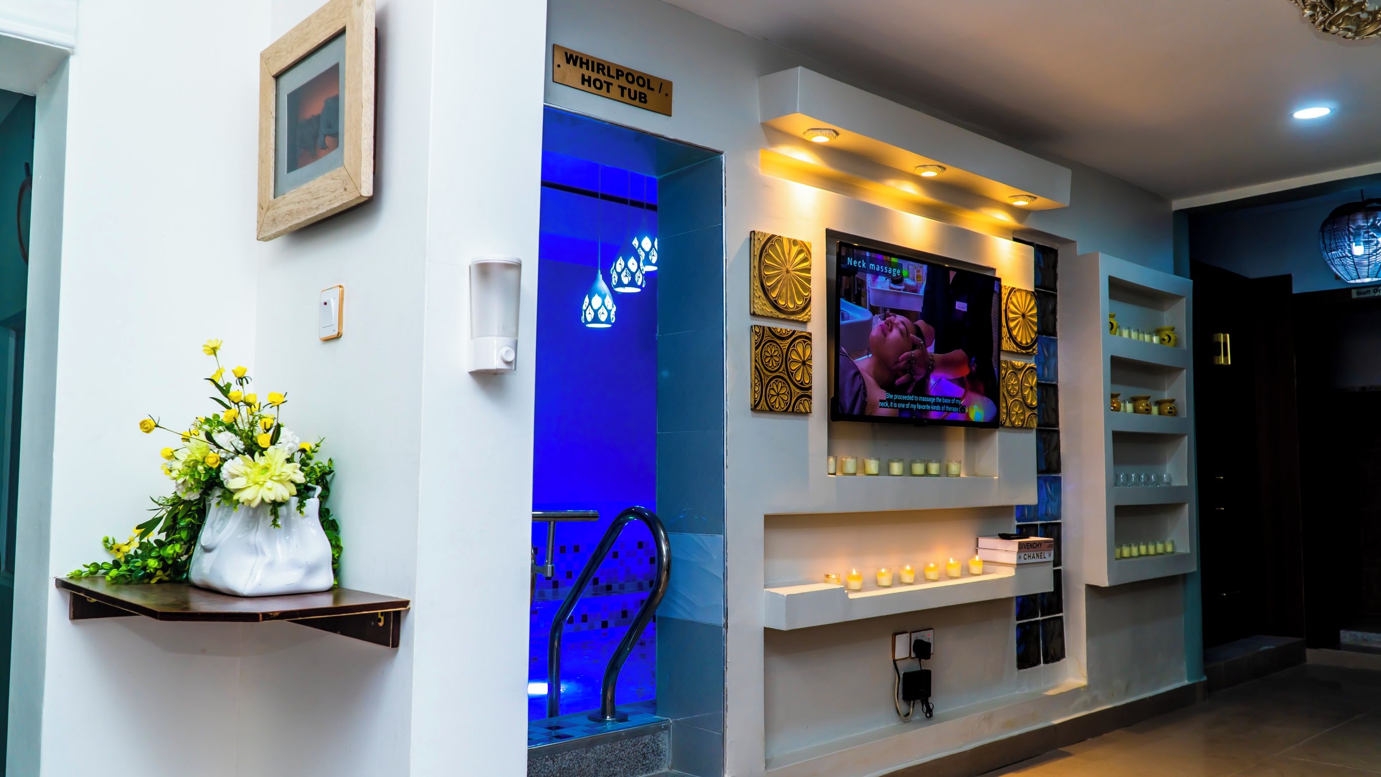 A reception area with a television displaying a massage service, a blue-lit entrance, and a shelf with candles.