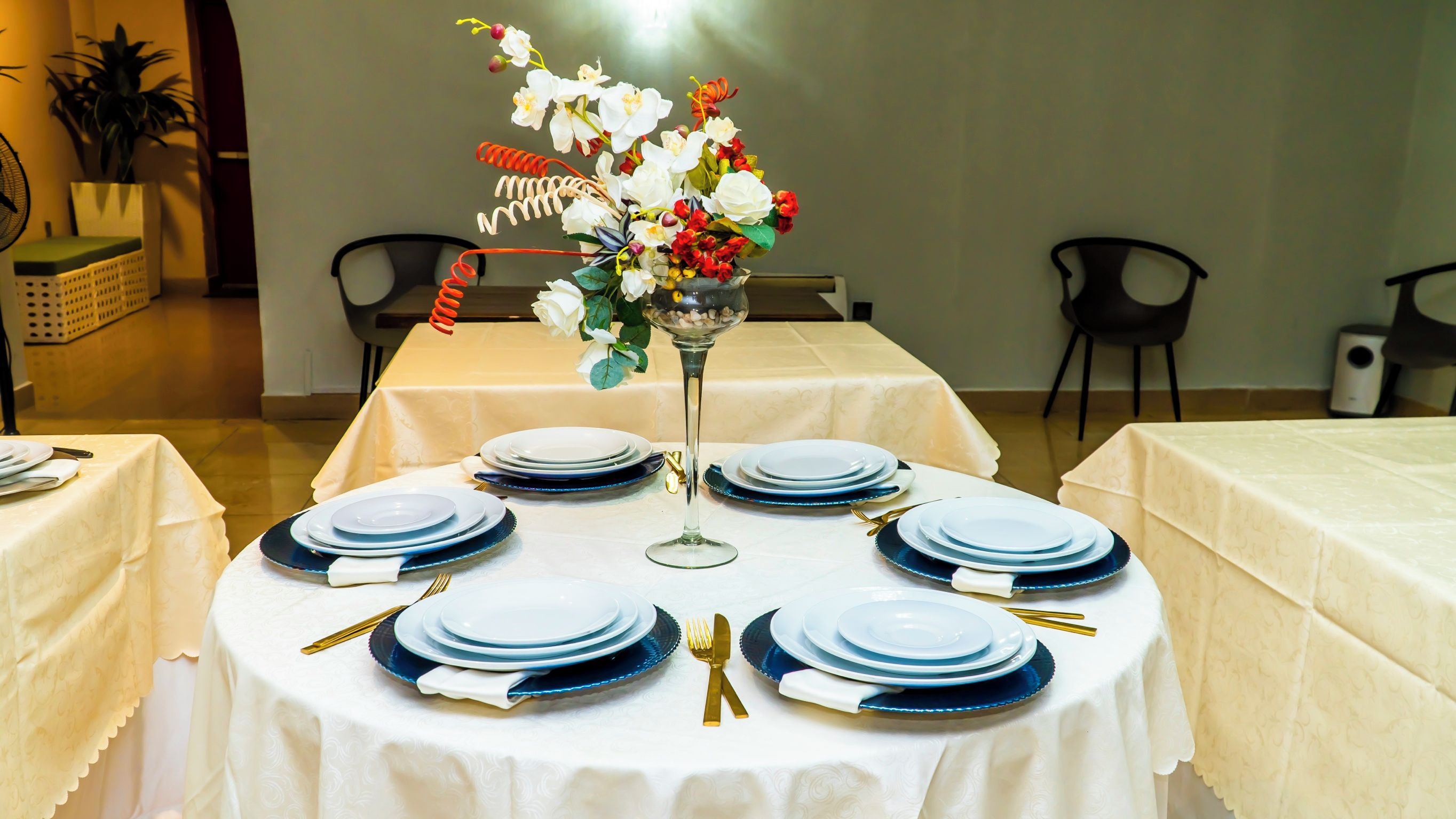 A round dining table with blue plates and gold cutlery, surrounded by chairs, in a room with a white tablecloth.