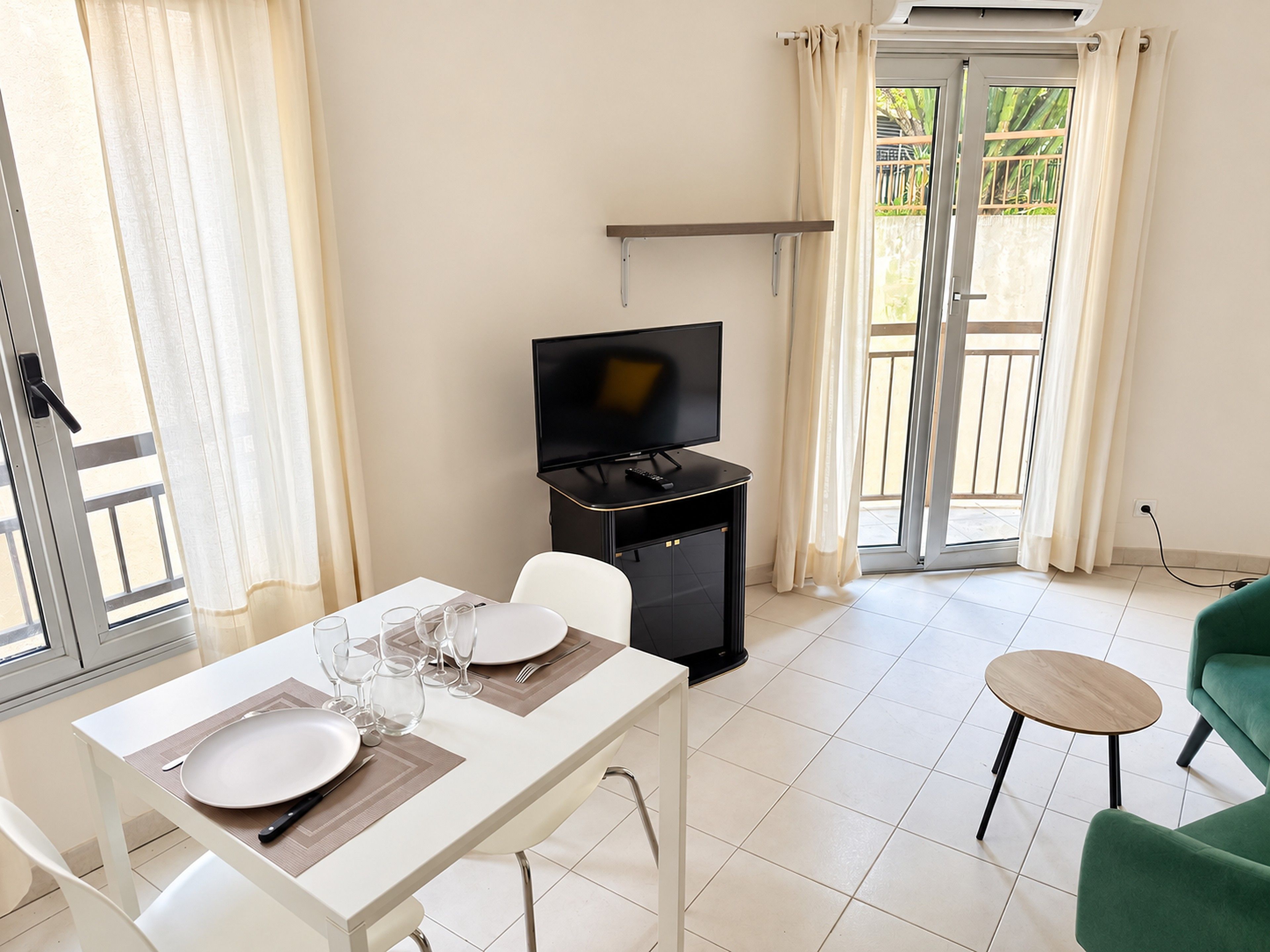 A modern living room with a dining table set for two, a flat-screen TV, and a sliding glass door leading to a balcony.