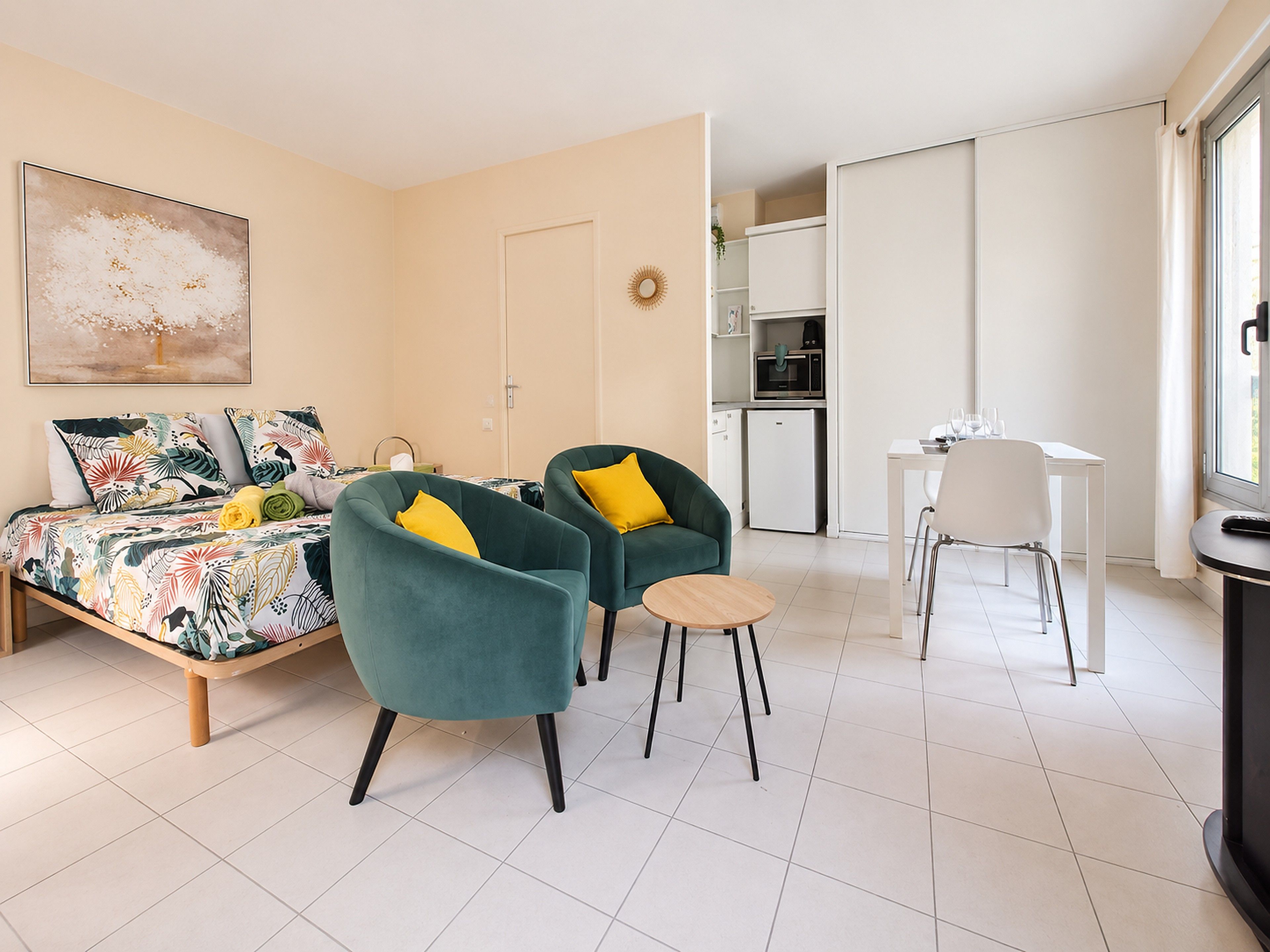 A modern, well-lit room with a bed, two armchairs, a small round table, a dining table with chairs, and a kitchenette.
