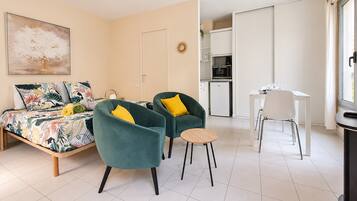 A modern, well-lit room with a bed, two armchairs, a small round table, a dining table with chairs, and a kitchenette.