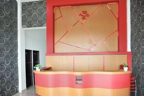 Un comptoir d’accueil avec un panneau décoratif aux motifs géométriques, deux plantes en pot et une porte portant un numéro.