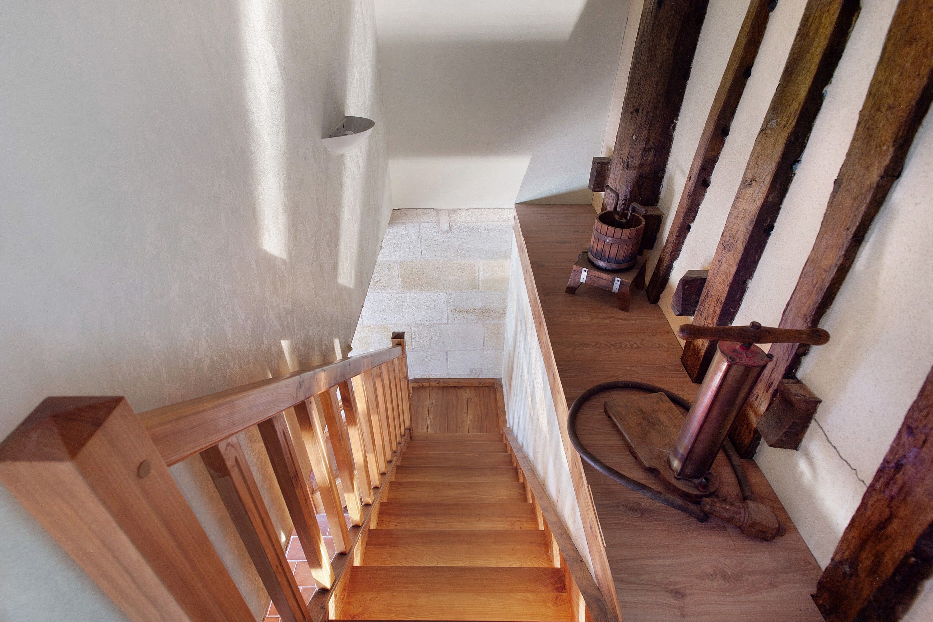 Una escalera de madera con barandilla que lleva a un rellano con pared blanca y un barril de madera.