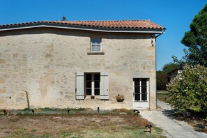 Un bâtiment en pierre de deux étages, avec un toit recouvert de tuiles rouges, deux fenêtres dotées de volets et un petit jardin à l’avant.