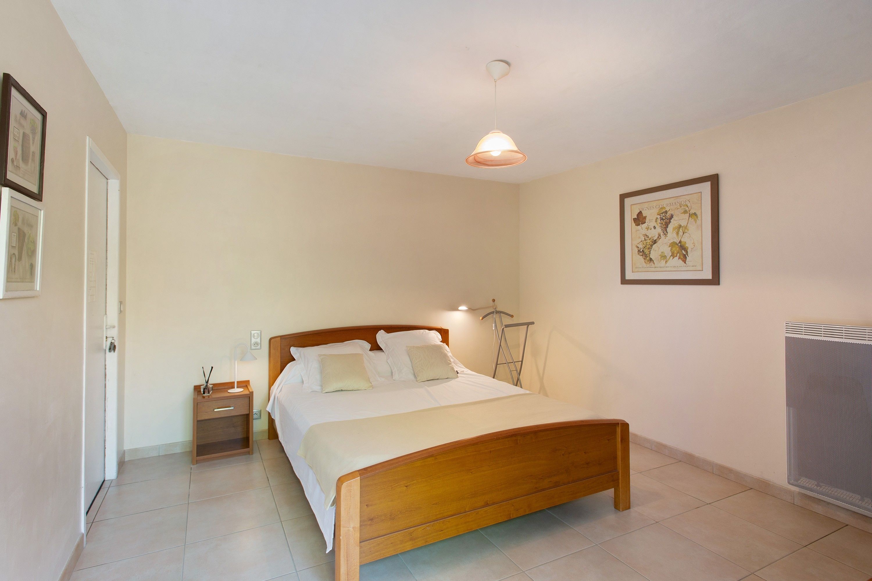 A bedroom with a wooden bed, bedside tables, a hanging lamp, a radiator, and framed artwork on the wall.