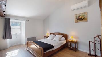 A bedroom with a wooden bed, two bedside tables with lamps, a window with curtains, and a wall-mounted air conditioner.