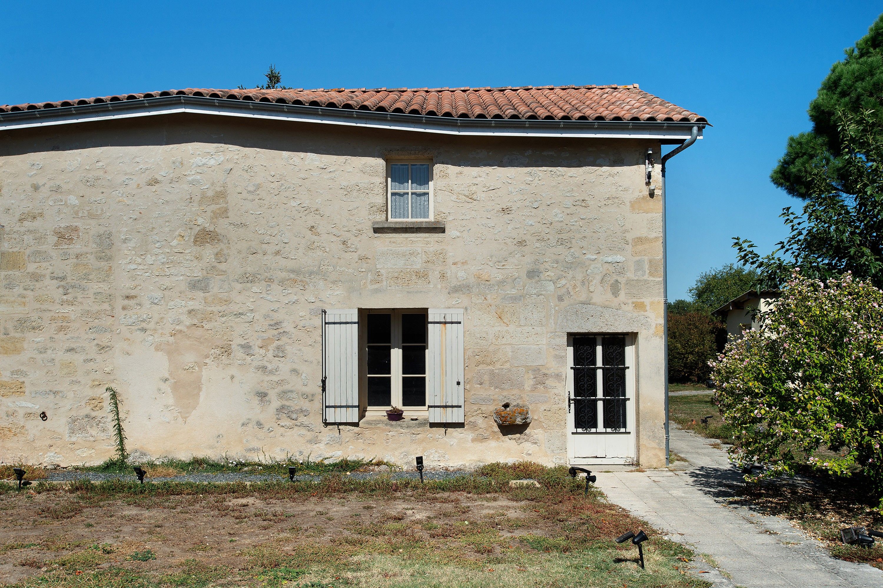 A two-story stone building with a red-tiled roof, two windows with shutters, and a small garden area in front.