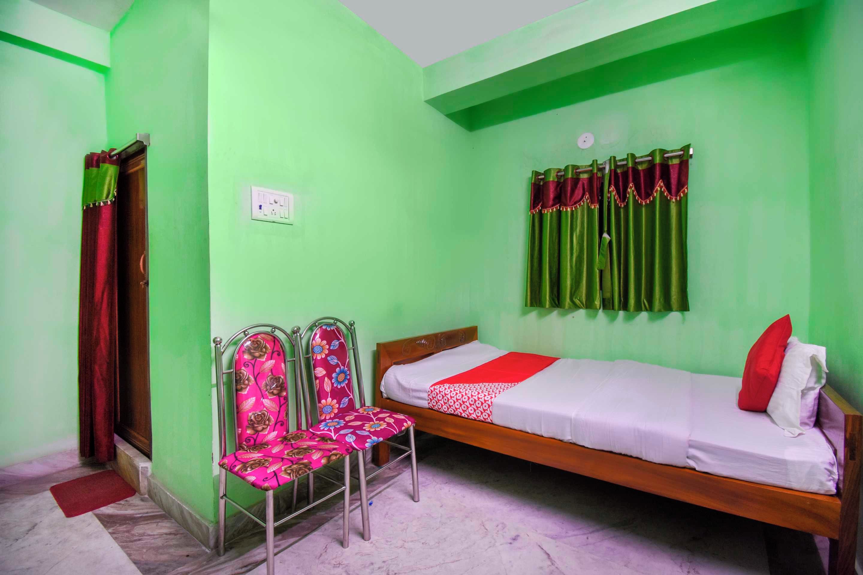 A small, single-bed room with a green wall, a wooden bed frame, a metal chair, and a red curtain.