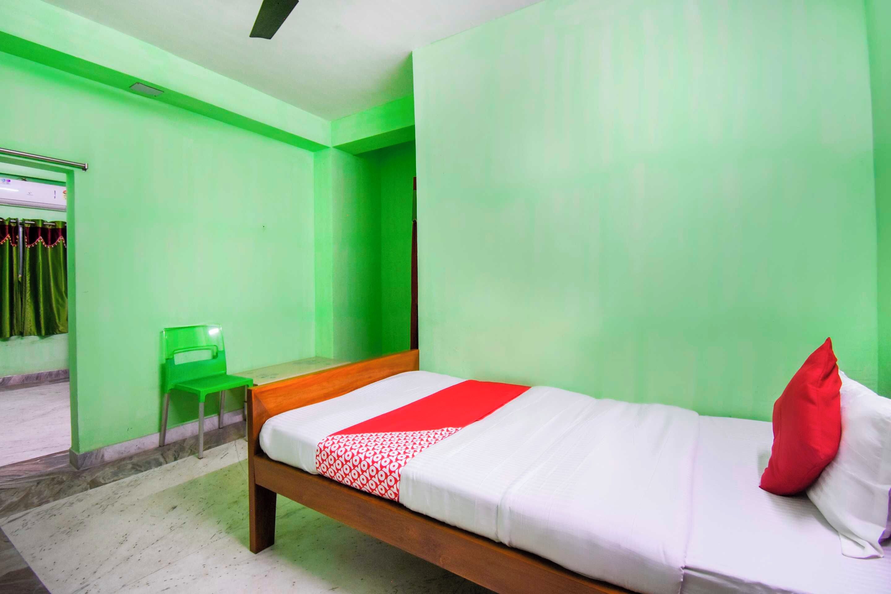A single bed with a wooden frame, white bedding, and a red pillow in a room with green walls and a green chair.