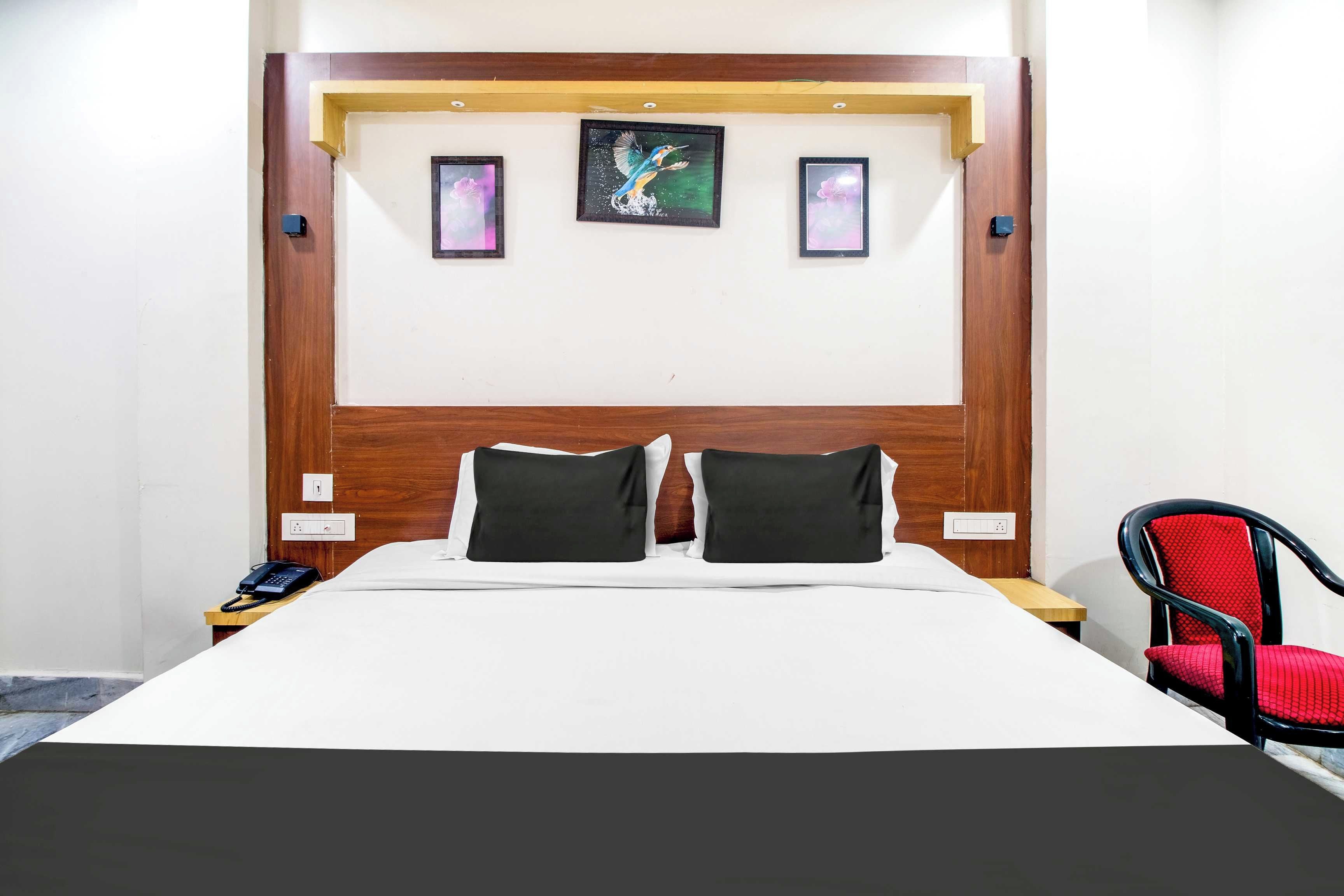 A hotel room with a wooden headboard, two black pillows, a white bedspread, and a single red chair.