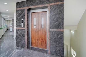 A hallway with a marble floor, a wooden elevator door, and a green counter. - Hotel O Magunta Layout Nellore (Nellore)