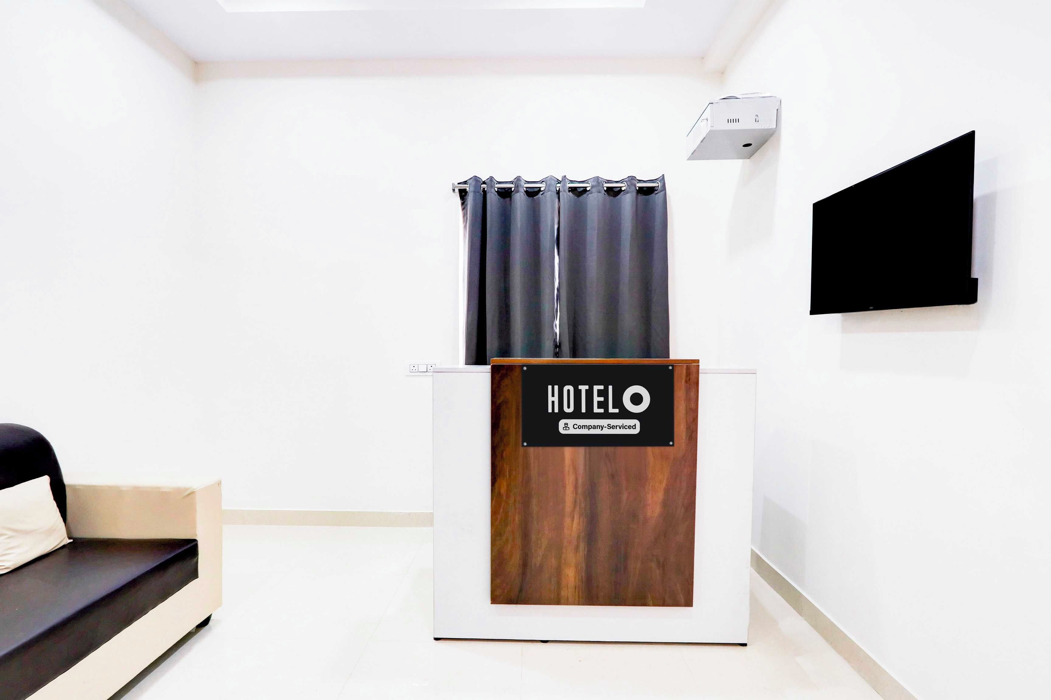 Uma área de recepção de hotel com uma mesa de madeira, uma placa indicando 'HOTELO Company Serviced', uma TV de tela plana preta e uma janela com cortinas.