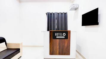 Uma área de recepção de hotel com uma mesa de madeira, uma placa indicando 'HOTELO Company Serviced', uma TV de tela plana preta e uma janela com cortinas.