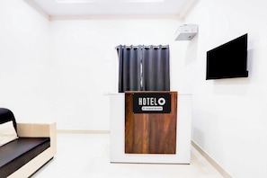 Zona de recepción de un hotel con un escritorio de madera, un letrero que dice 'HOTELO Company Serviced', un televisor de pantalla plana negro y una ventana con cortinas.