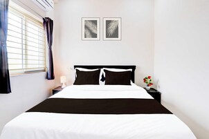 Una habitación de hotel con una cama, una mesita de noche, una lámpara, una ventana con cortinas y dos adornos con motivos de hojas en la pared.