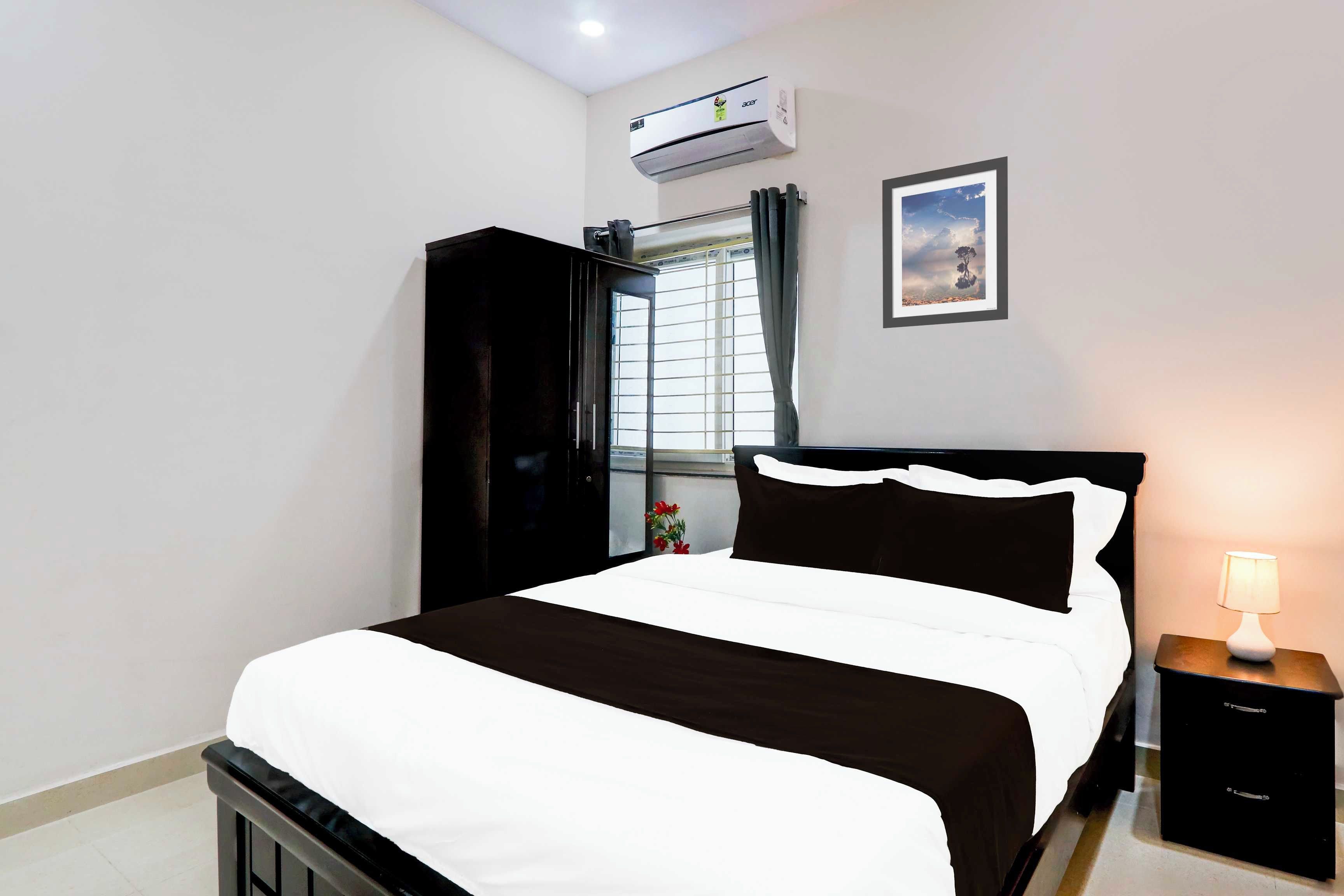 Un dormitorio con cama, mesita de noche, lámpara, equipo de aire acondicionado, ventana con cortinas e imagen enmarcada en la pared.